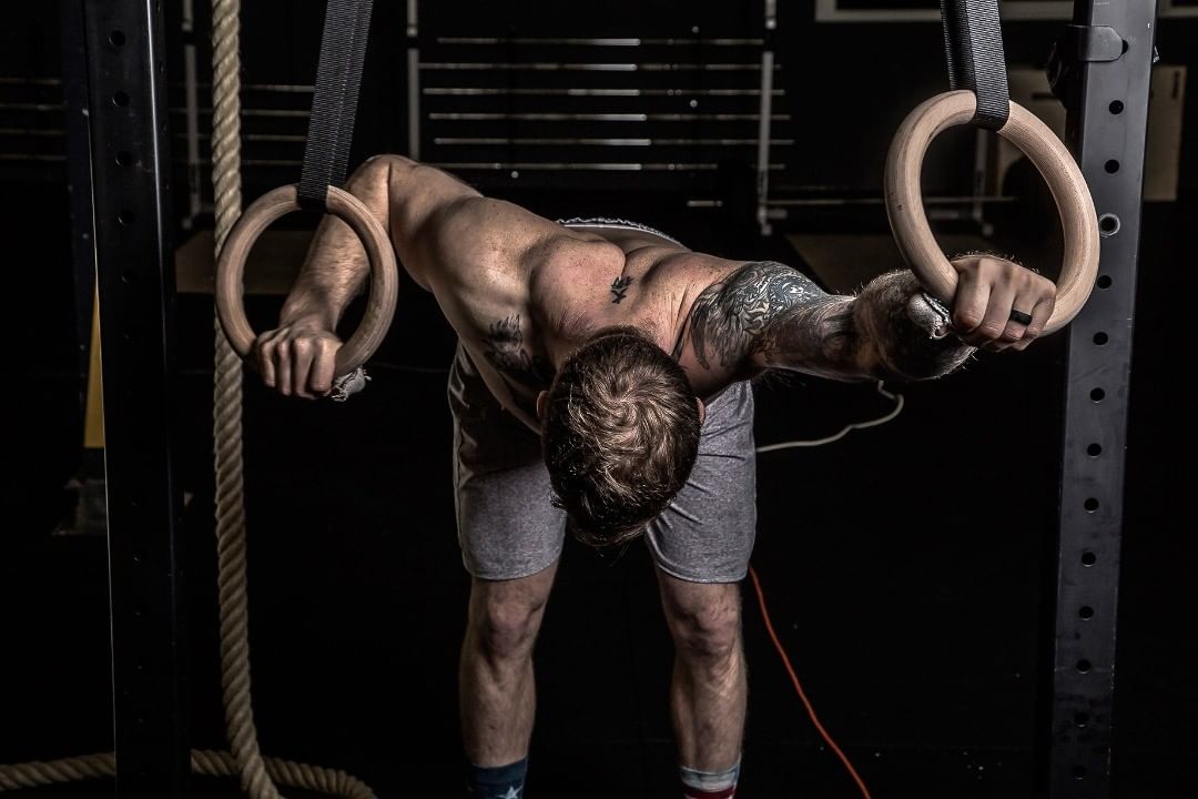 Muscular athlete hanging forward from gymnastic rings, head down, in a darkened training hall — sports photograph by Hamudi Leon