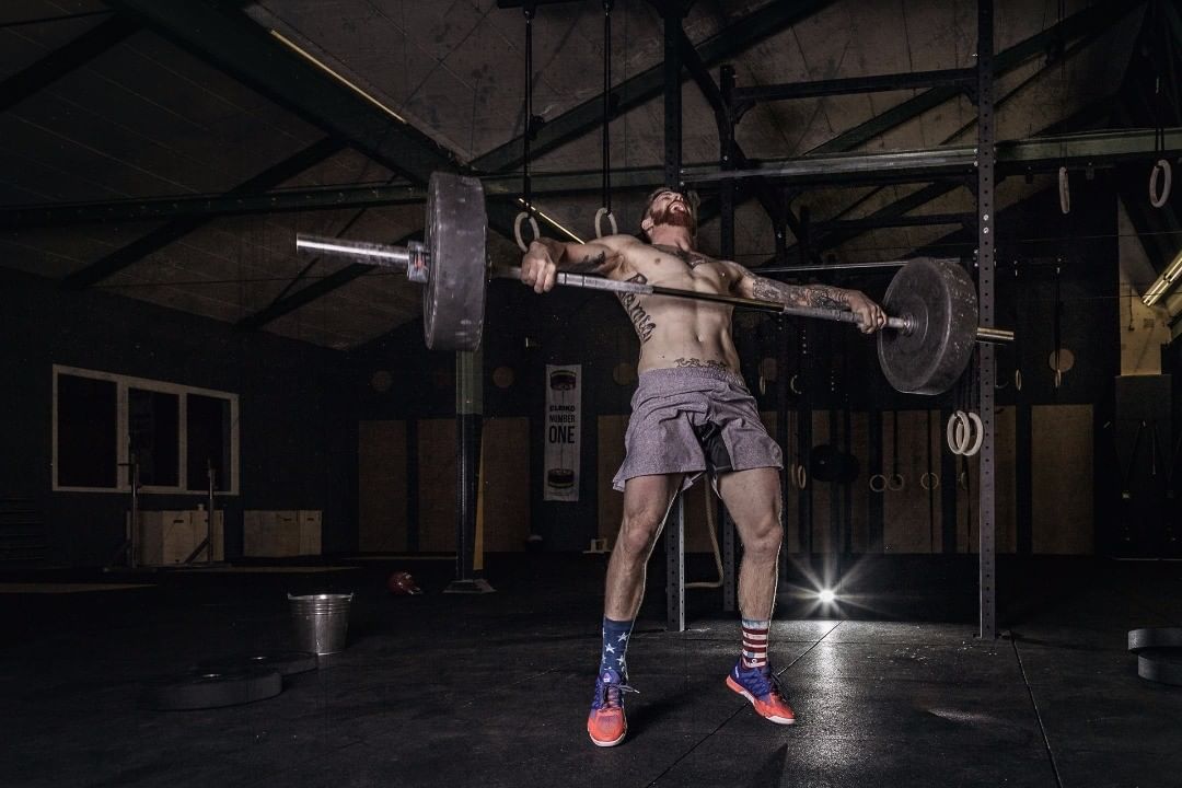 Athlete locking out a loaded barbell overhead in a dim industrial gym — sports photograph by Hamudi Leon