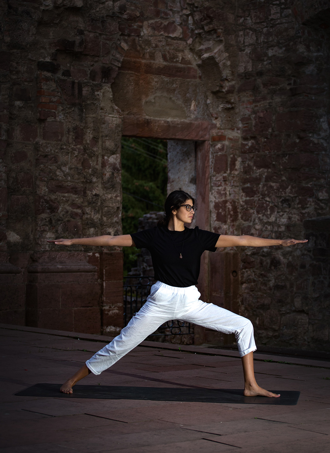 Woman in warrior pose on a mat before the arched doorway of a stone ruin — sports photograph by Hamudi Leon