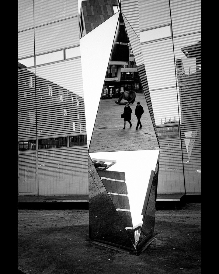Angular mirrored sculpture on a square, reflecting a facade and two passers-by — street photograph by Hamudi Leon