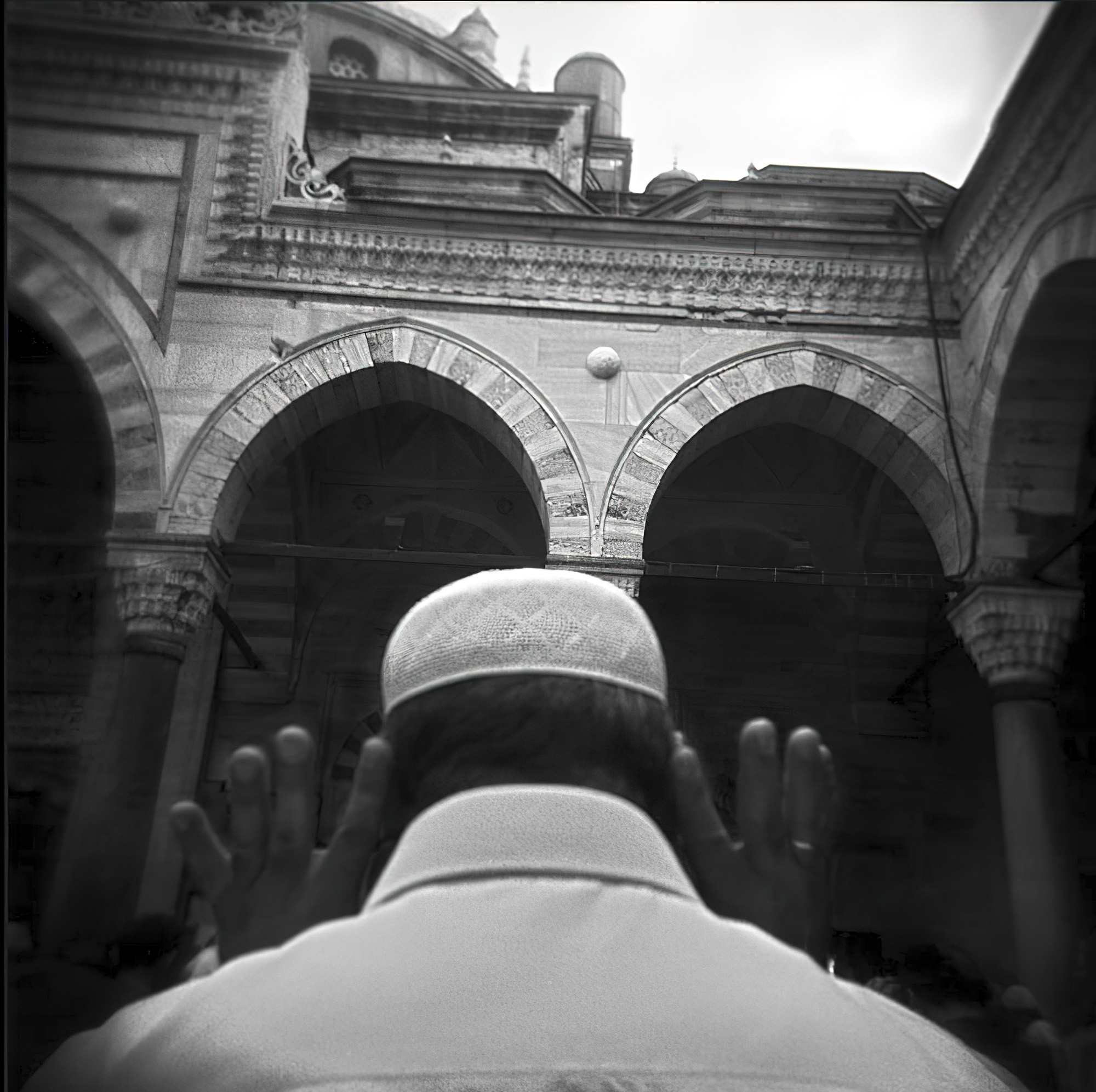 Man in a white cap seen from behind with his hands raised, facing an arcaded courtyard — street photograph by Hamudi Leon