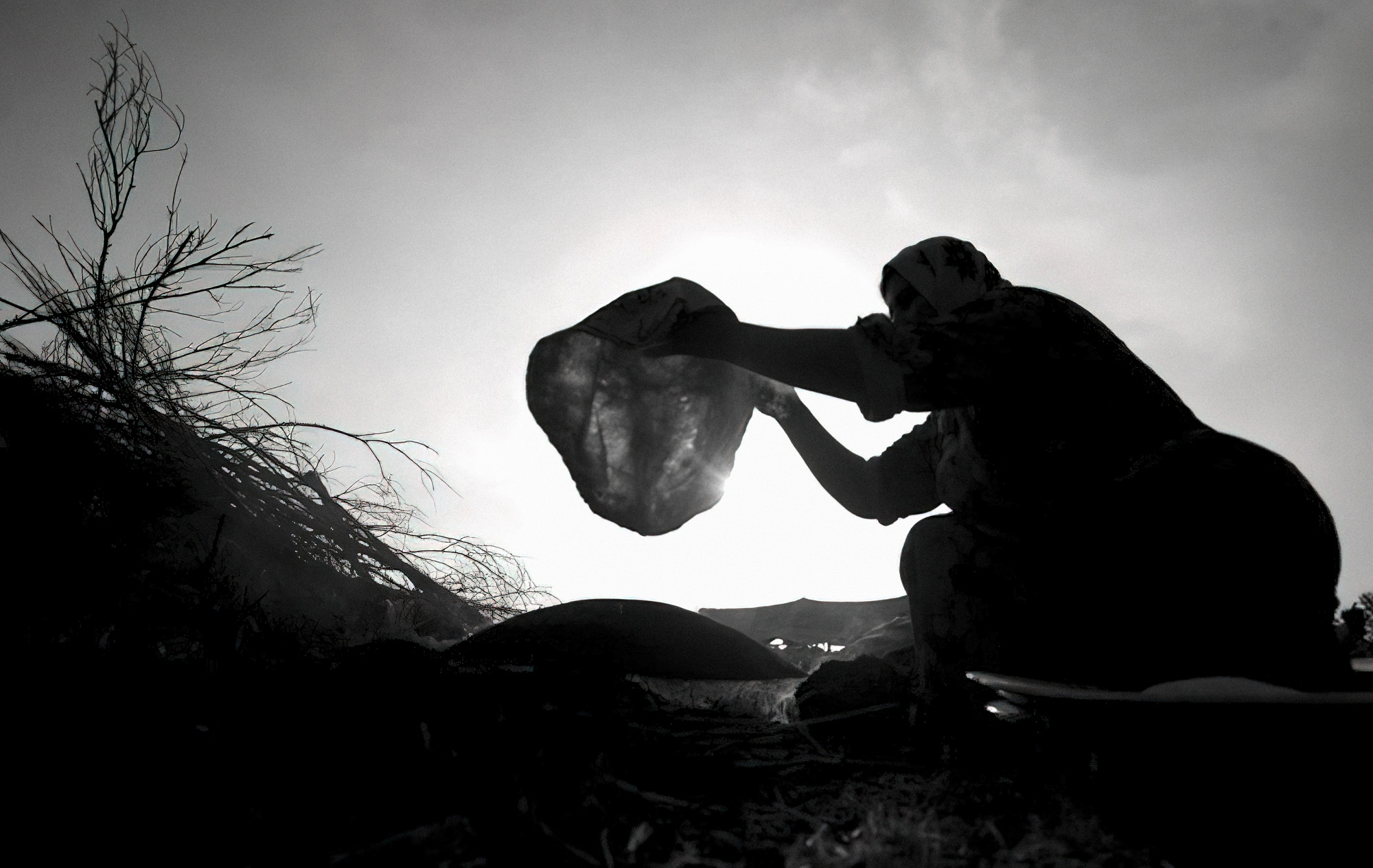 Silhouette of a person lifting a bundle against a blinding sky beside a bare tree — street photograph by Hamudi Leon