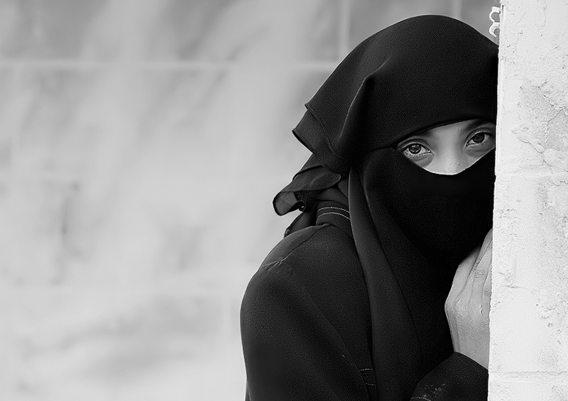 Woman in a niqab, only her eyes uncovered, against a pale wall — street photograph by Hamudi Leon