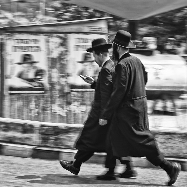 Two men in long coats and wide hats striding past a wall of posters, caught in motion blur — street photograph by Hamudi Leon