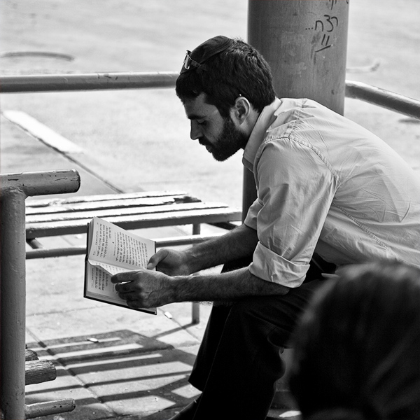 Bearded man in a white shirt reading a book at an outdoor bench — street photograph by Hamudi Leon