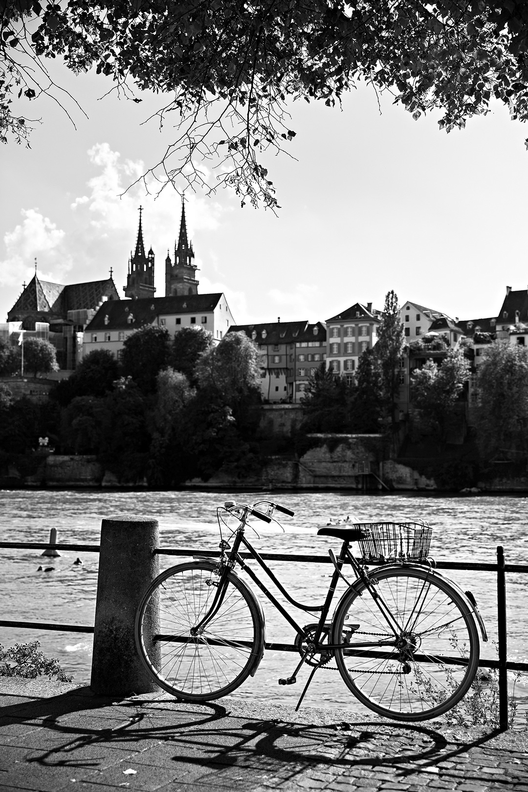 Bicycle leaning on a railing above a river, the old town and its church behind — street photograph by Hamudi Leon