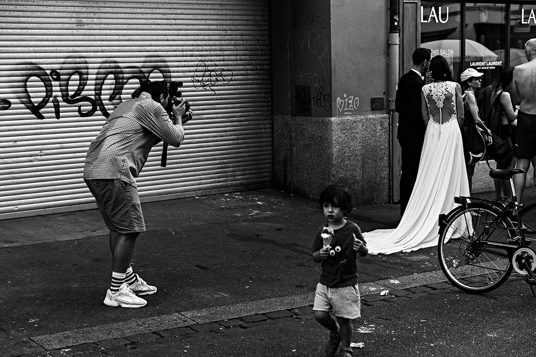 Photographer crouching to shoot a bride while a small child wanders past a shuttered, graffitied front — street photograph by Hamudi Leon