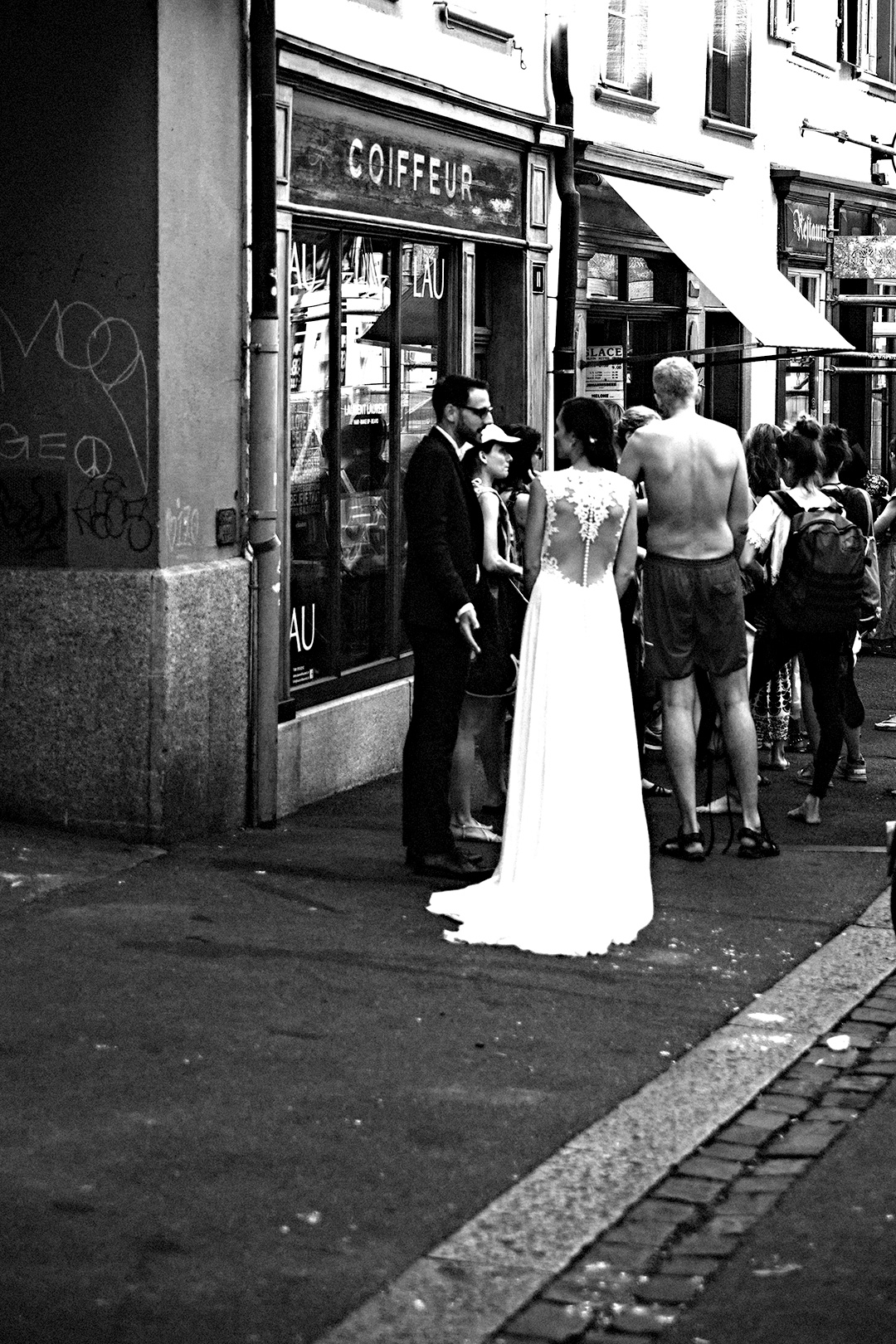Bride and groom on a crowded pavement outside a hairdresser's window — street photograph by Hamudi Leon