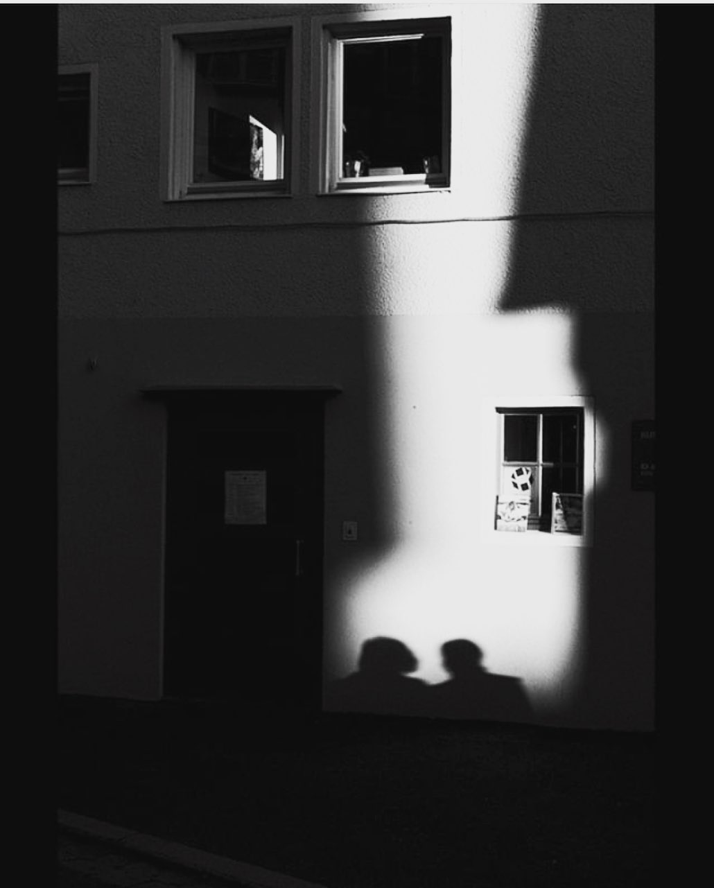 Two shadows thrown across a plain facade by a hard diagonal of afternoon light — street photograph by Hamudi Leon