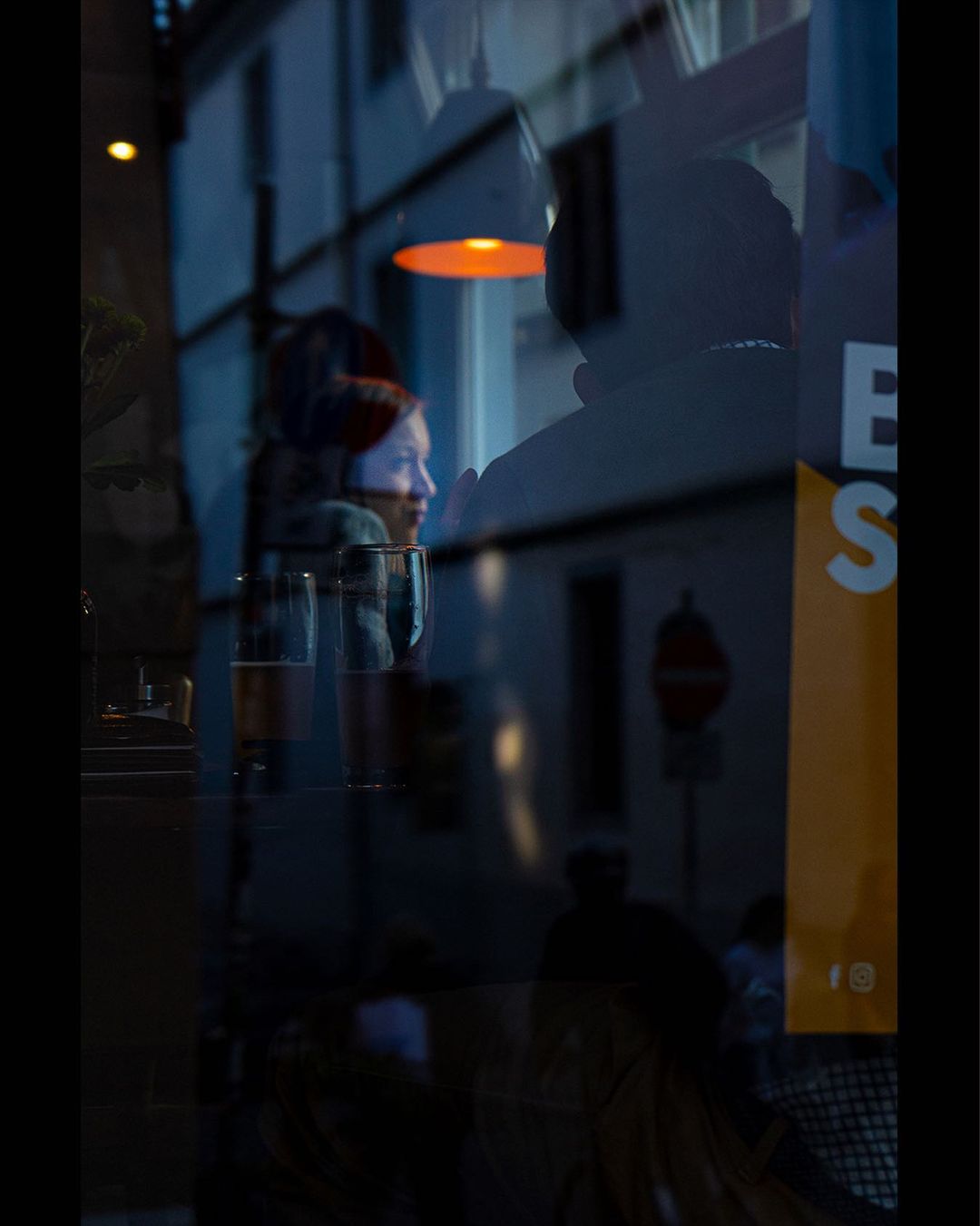 Couple in a cafe seen through blue-lit glass and its reflections — street photograph by Hamudi Leon
