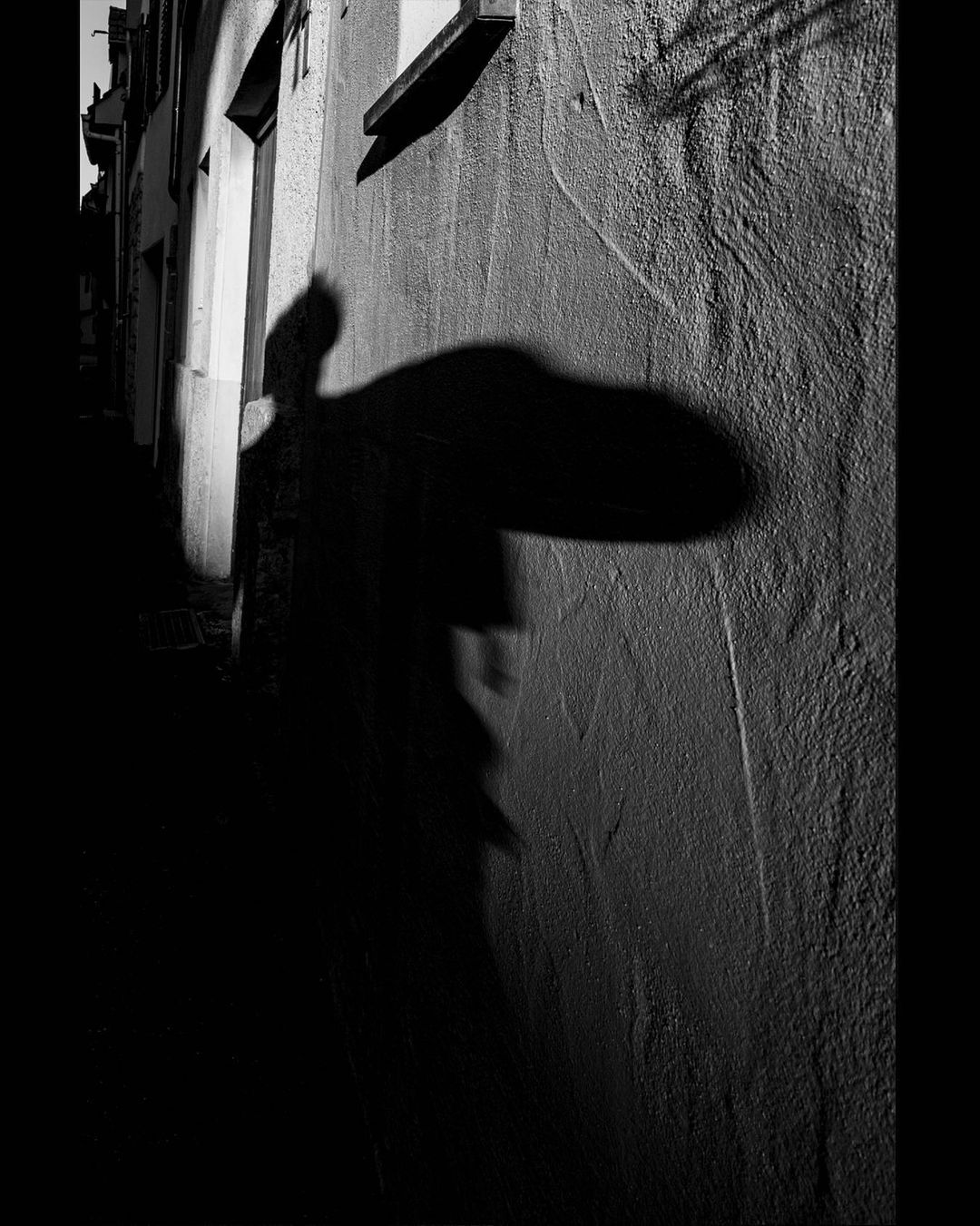 Shadow of a person in a wide hat thrown across a rough plastered alley wall — street photograph by Hamudi Leon