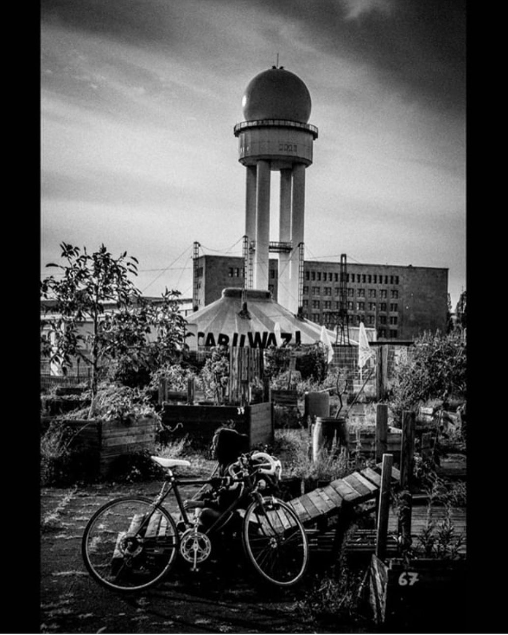 Bicycles parked before a disused airport control tower and allotment gardens — street photograph by Hamudi Leon