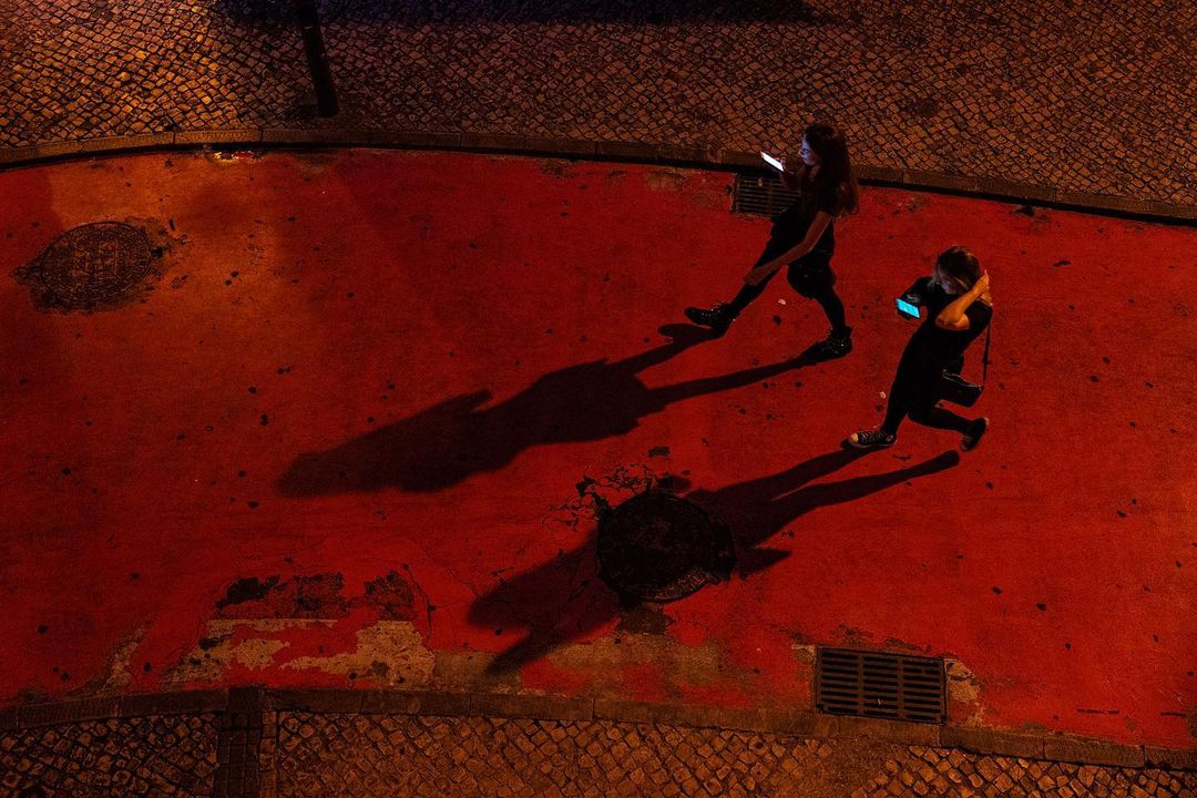 Two people crossing a red-lit square at night, their shadows stretched far ahead — street photograph by Hamudi Leon
