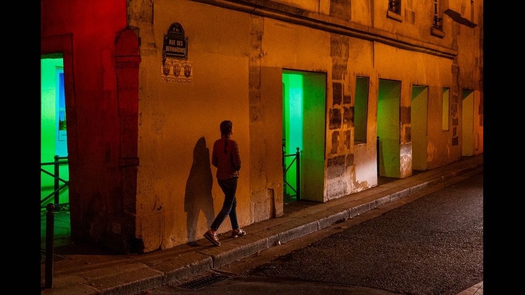 Person walking an alley at night between green and orange shopfront light — street photograph by Hamudi Leon