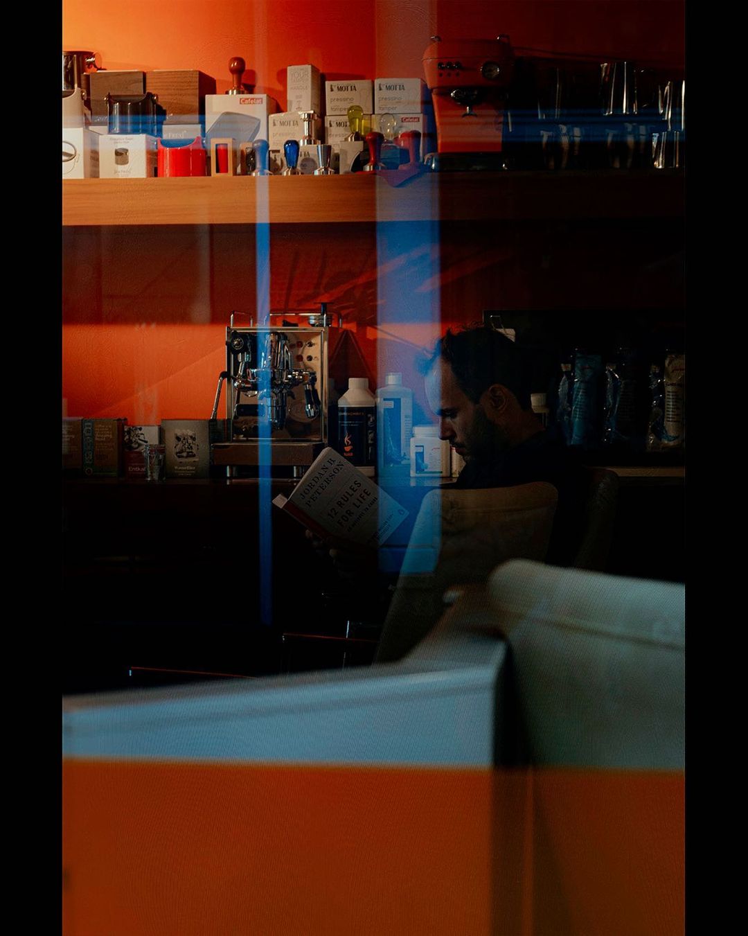 Man reading a newspaper inside a shop, seen through orange and blue reflected glass — street photograph by Hamudi Leon
