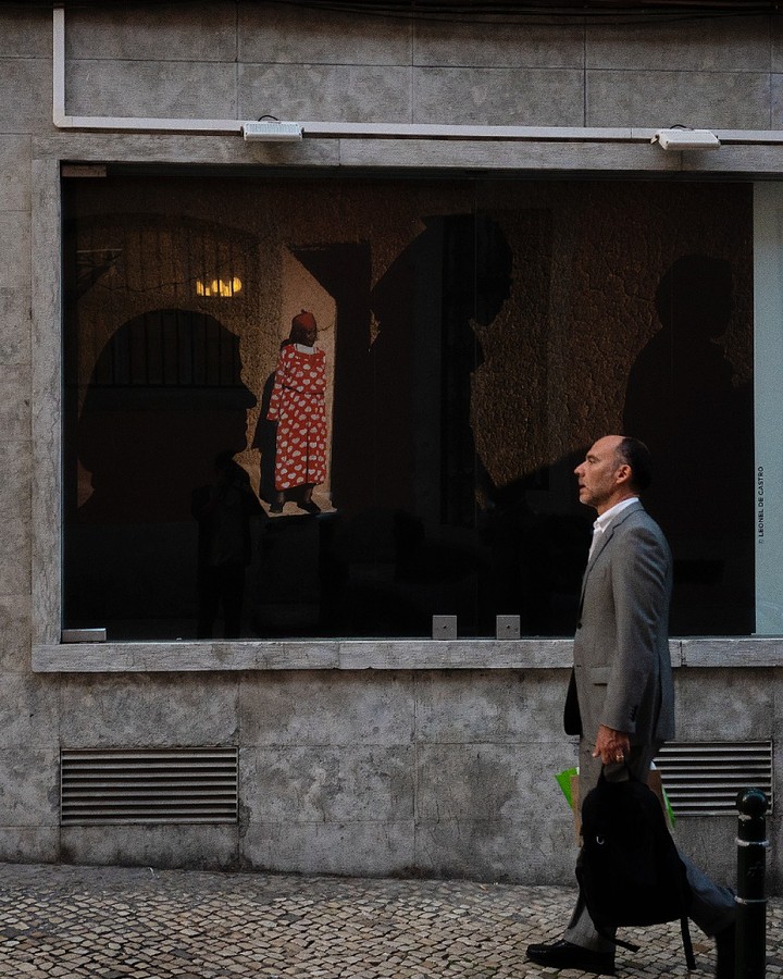 Man in a suit passing a shop window whose display holds a red-patterned figure — street photograph by Hamudi Leon