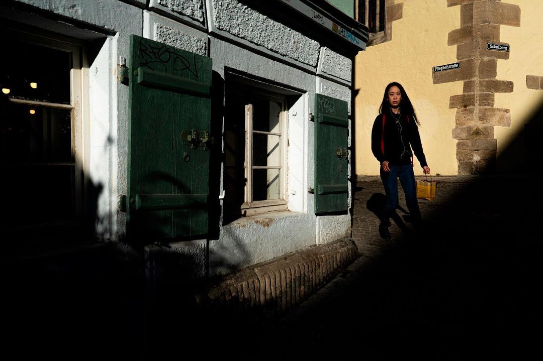 Woman stepping into sunlight at the end of a shaded alley of green shutters — street photograph by Hamudi Leon