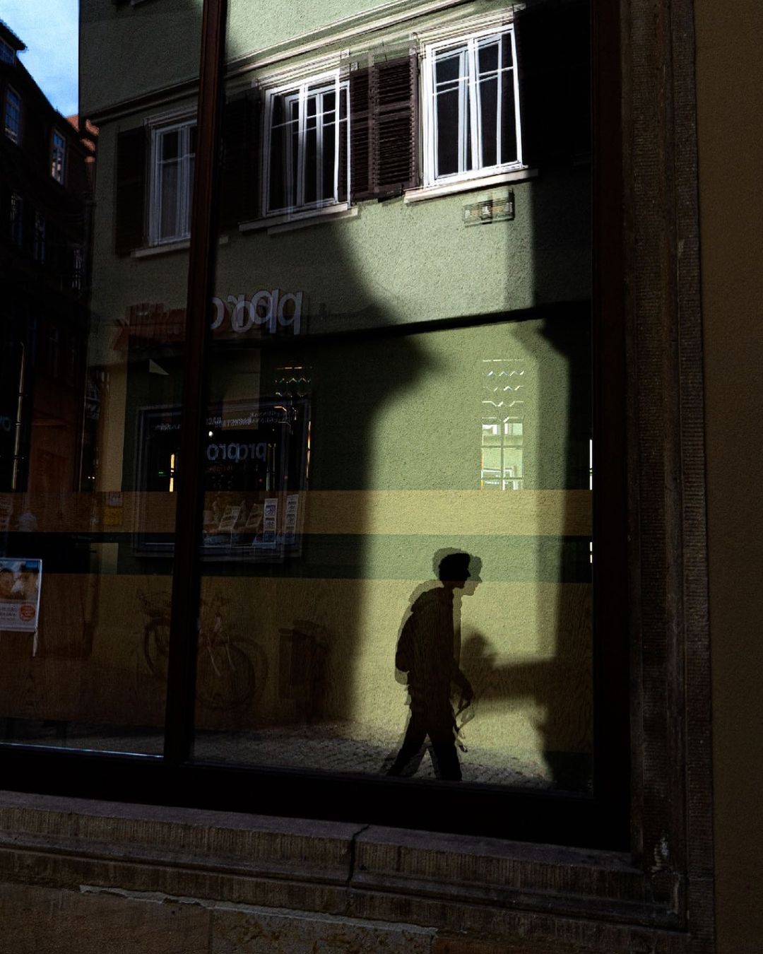 Silhouette of a walker reflected in a large window with a green facade behind — street photograph by Hamudi Leon