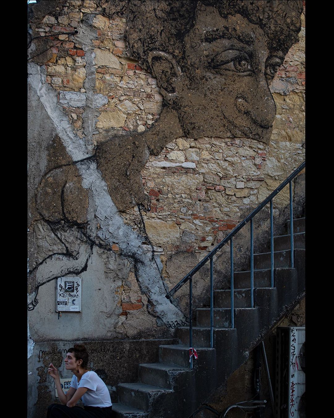 Person sitting on a step beneath a huge charcoal face painted on a cracked wall — street photograph by Hamudi Leon