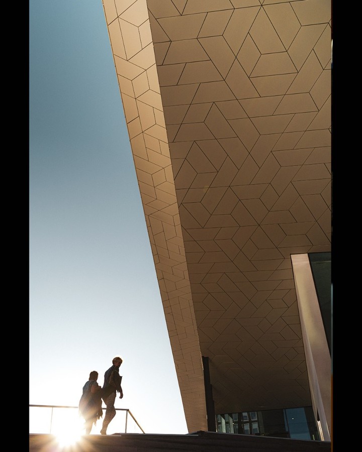 Two silhouettes on a stair against the sun, under a patterned overhang — street photograph by Hamudi Leon