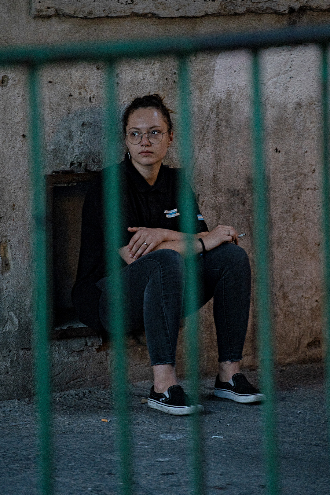 Young woman sitting against a weathered wall, seen through green railings — street photograph by Hamudi Leon