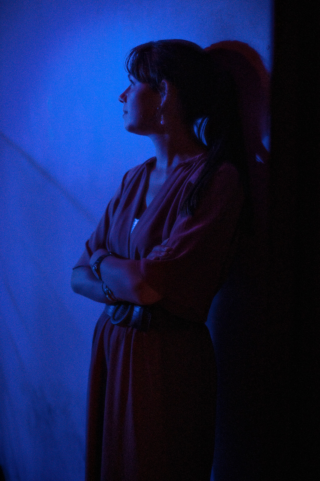 Woman in profile against a deep blue wall, arms folded — street photograph by Hamudi Leon