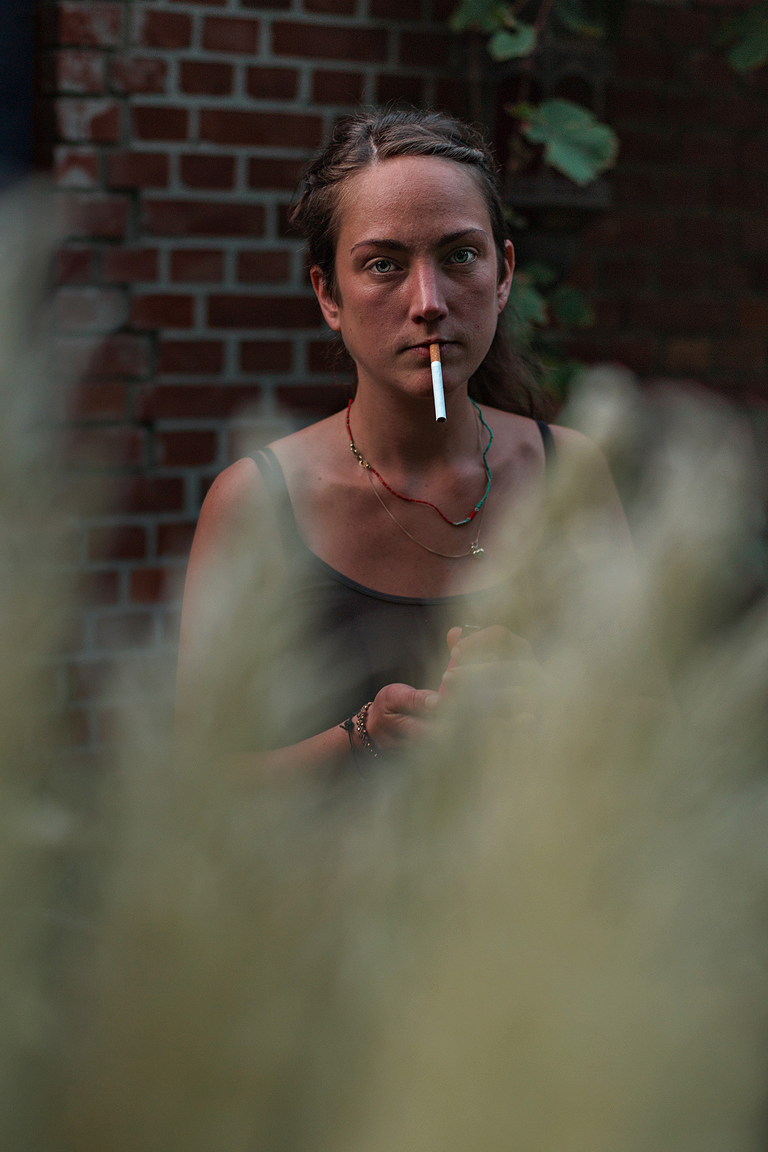 Woman smoking outside a brick building, seen past out-of-focus grass — street photograph by Hamudi Leon