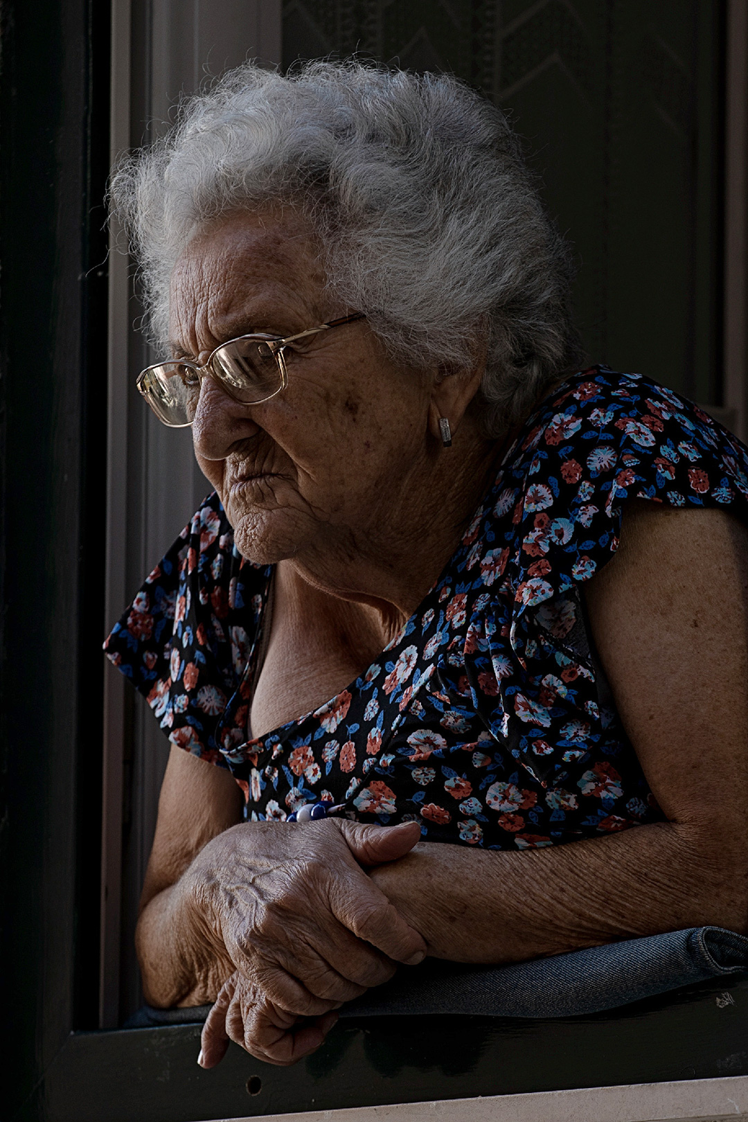 Elderly woman leaning on a windowsill, looking out into the street — street photograph by Hamudi Leon