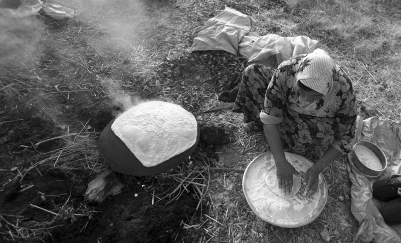 Woman baking flatbread over an open fire on the ground — street photograph by Hamudi Leon