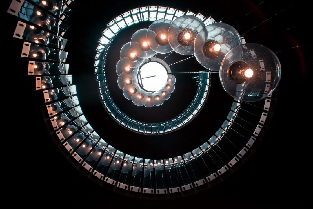 Spiral staircase seen from below, ringed with lamps — street photograph by Hamudi Leon