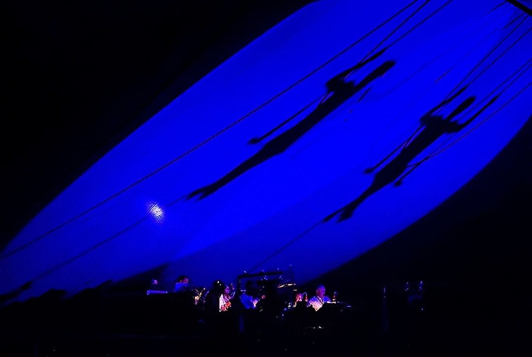 Silhouettes on a blue-lit stage canopy above an orchestra — street photograph by Hamudi Leon