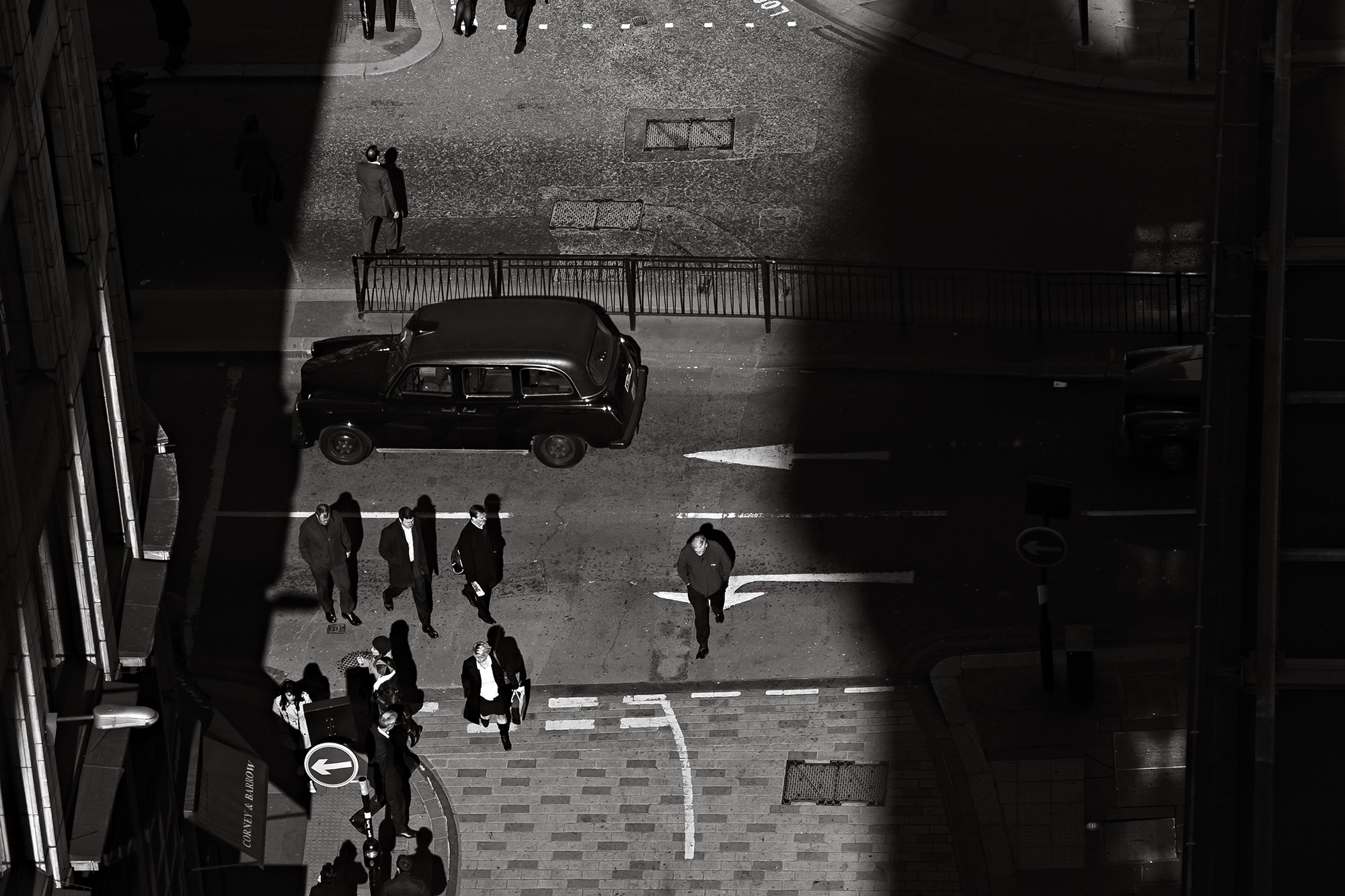 Street crossing seen from high above, a black taxi and pedestrians in hard light — street photograph by Hamudi Leon