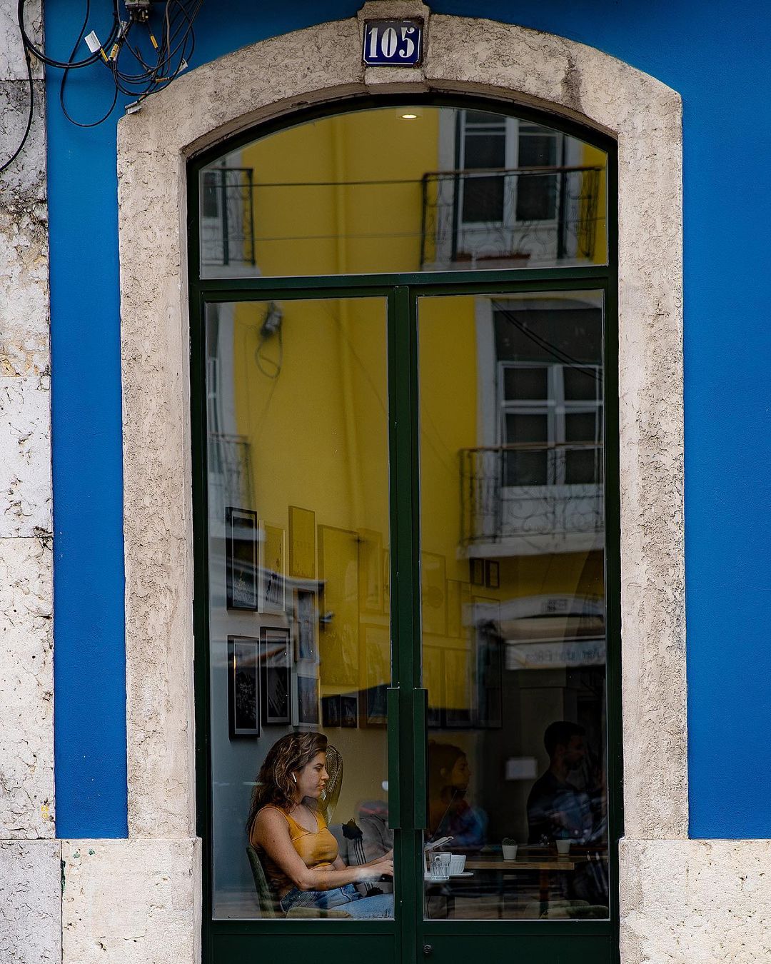 Woman working at a laptop inside a cafe, framed by a blue and yellow doorway — street photograph by Hamudi Leon