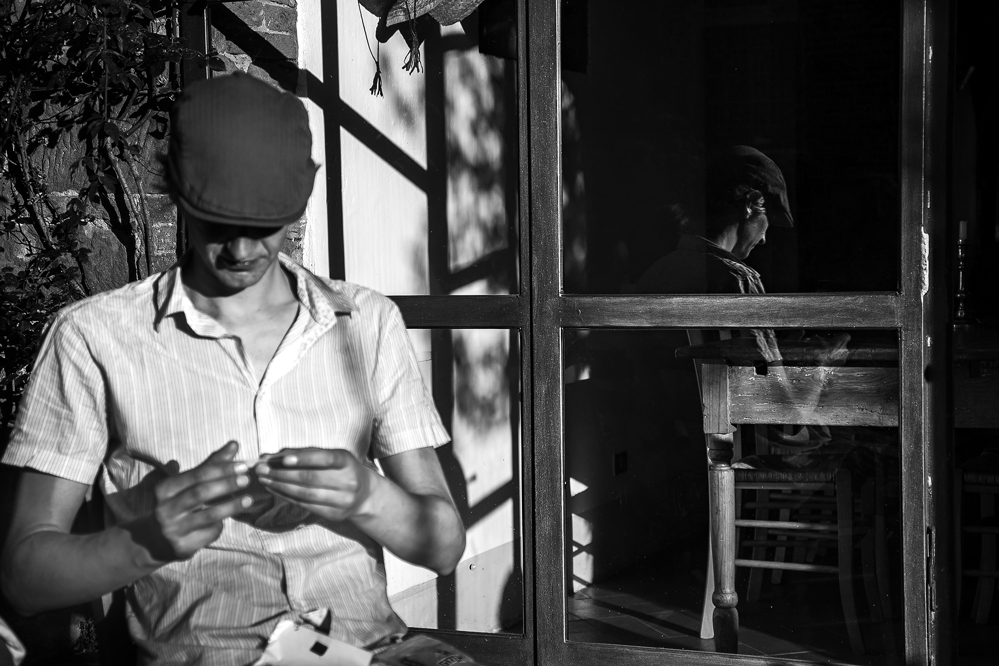 Man in a flat cap at a window, his reflection beside him in the glass — street photograph by Hamudi Leon