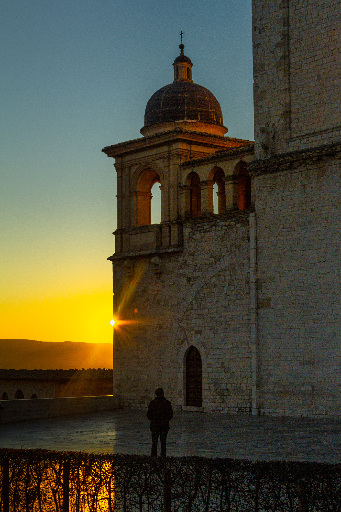 Church tower with a figure standing at the parapet as the sun sets behind the hills — street photograph by Hamudi Leon