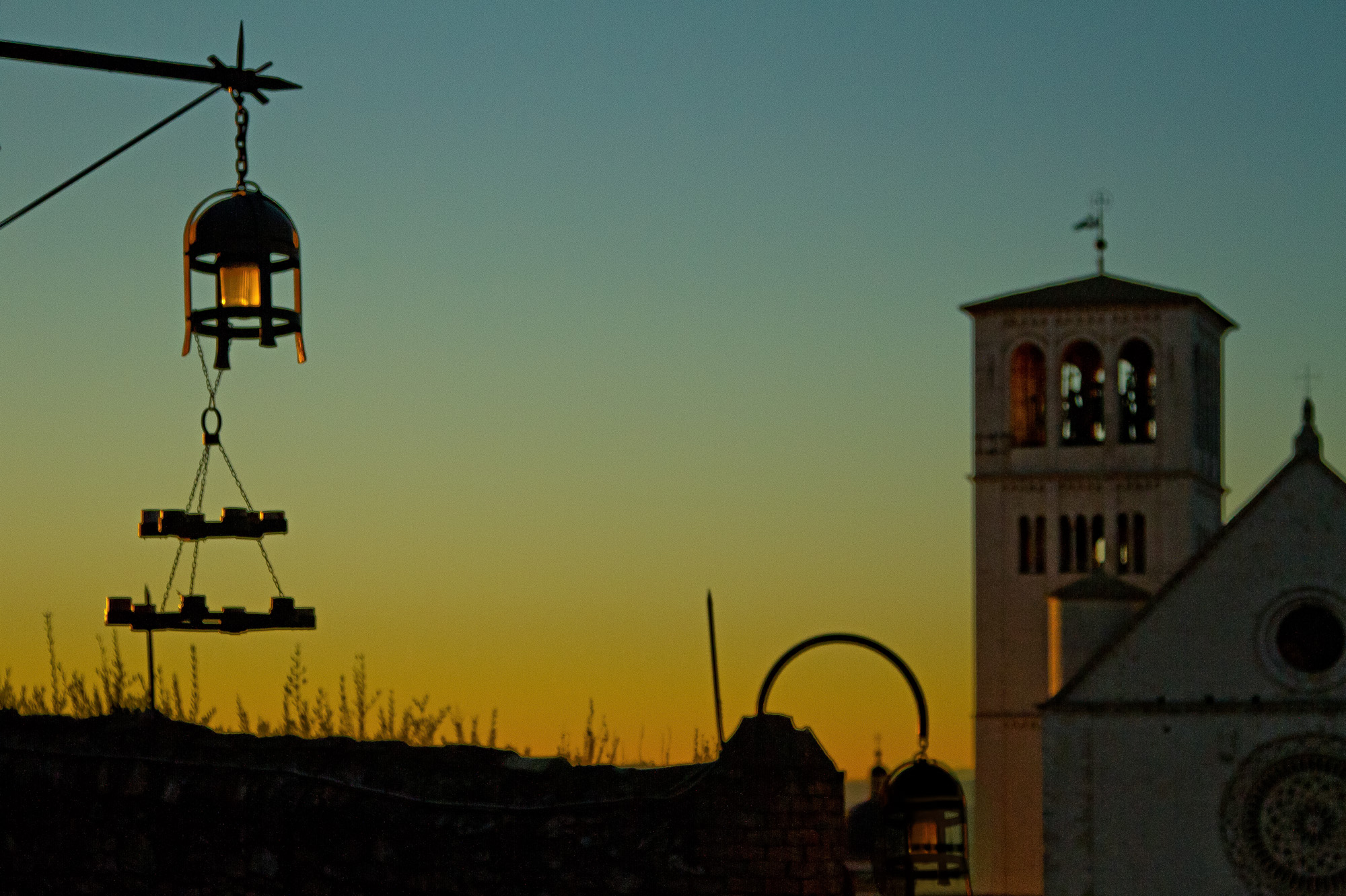 Wrought-iron lanterns hanging against a green evening sky beside a church tower — street photograph by Hamudi Leon