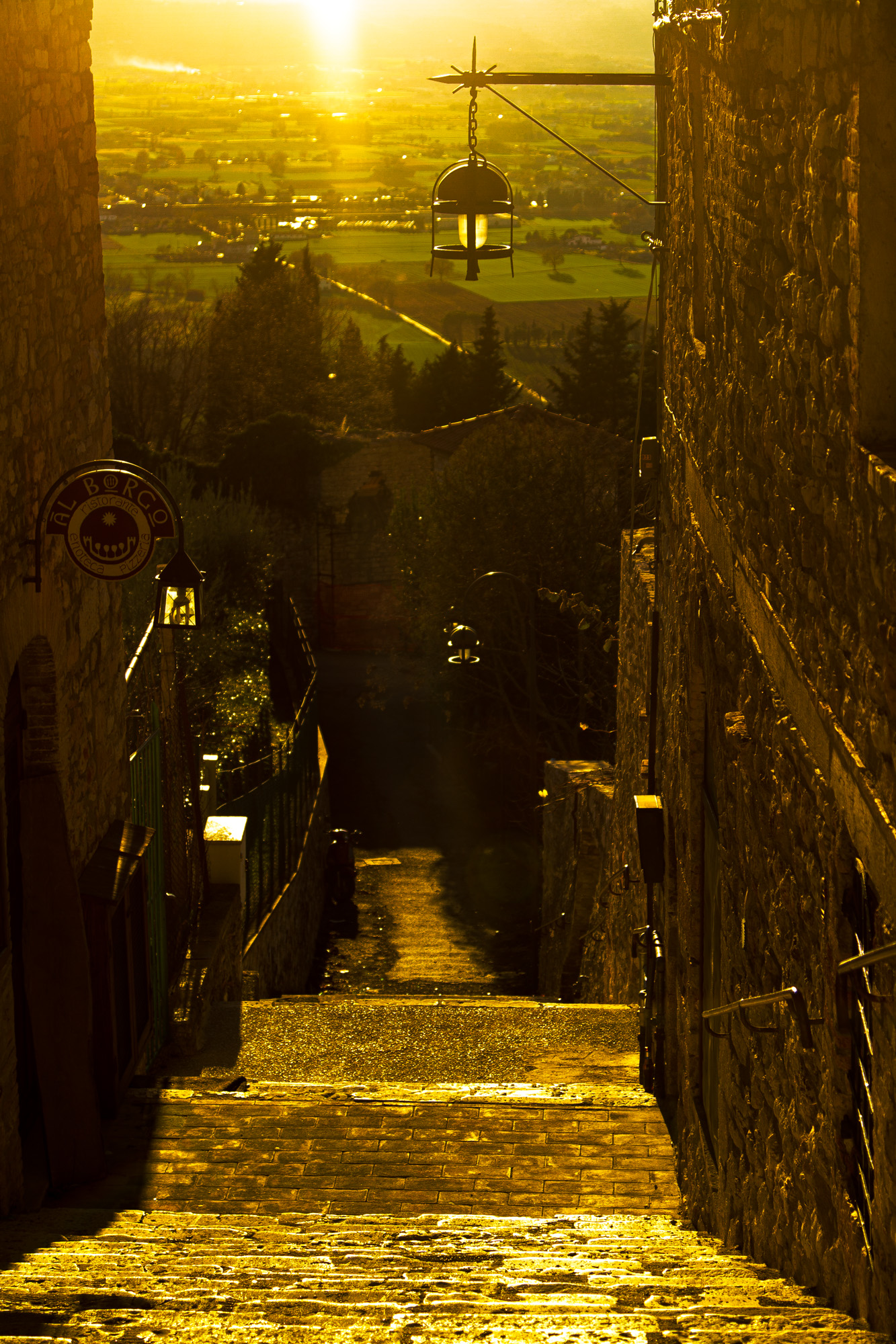 Wet cobbled stairway dropping steeply between stone houses toward a lit valley — street photograph by Hamudi Leon