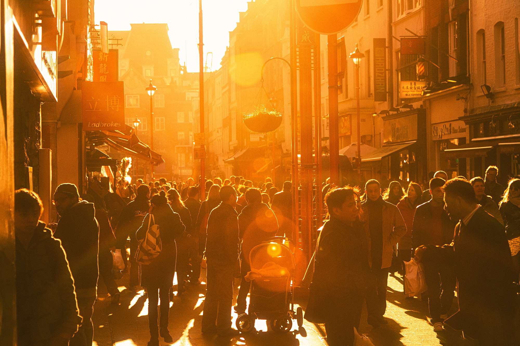 Crowd filling a narrow street, backlit by low sun — street photograph by Hamudi Leon